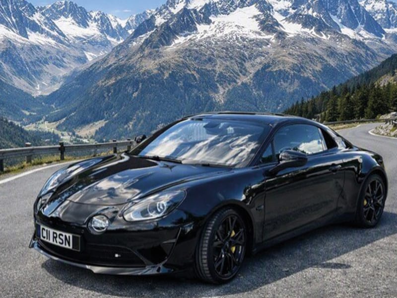 Black Alpine A110 parked on a winding mountain road, shown from the front three-quarter angle with snow-capped mountains in the background and front UK registration plate “C11 RSN.”