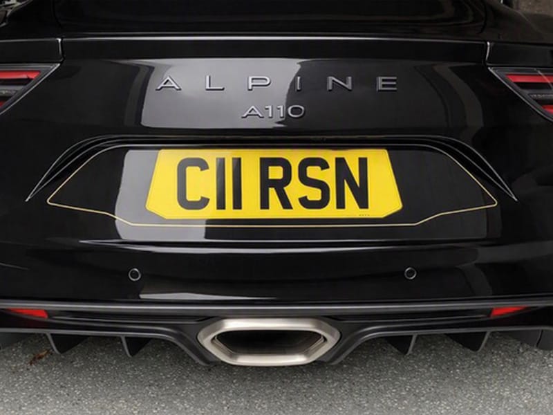 Close-up rear view of a black Alpine A110, featuring Alpine and A110 badging, a yellow shaped UK registration plate “C11 RSN,” parking sensors, central exhaust and rear diffuser.