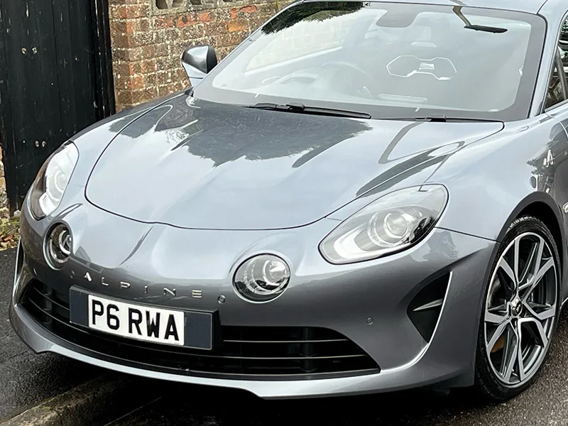 Grey Alpine sports car viewed from the front three-quarter angle, showing its rounded headlights, Alpine bonnet badging, alloy wheel and UK registration plate “P6 RWA.”