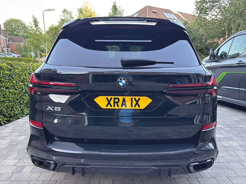 Rear view of a black BMW X5, showing slim LED taillights, twin exhaust surrounds and a yellow shaped Signature number plate displaying “XRA 1X.”