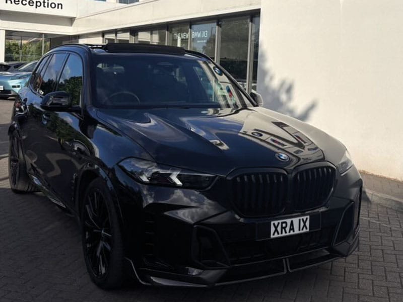 Front three-quarter view of a black BMW X5 parked outside a BMW dealership, featuring black exterior trim, dark alloy wheels and a white “XRA 1X” number plate.