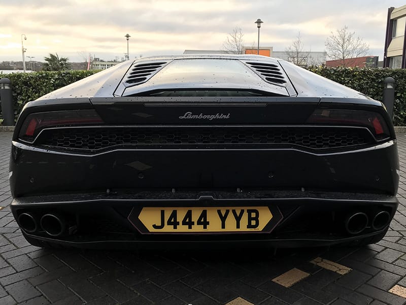 Straight rear view of a black Lamborghini Huracán, featuring the rear grille, quad exhausts and yellow shaped number plate “J444 YYB.”