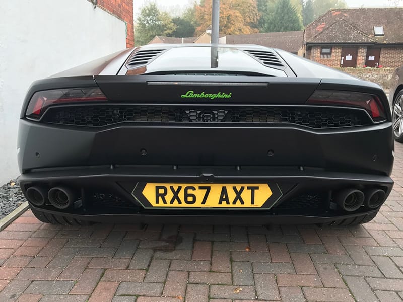 Rear view of a black Lamborghini Huracán, showing the rear grille, quad exhausts and a yellow shaped number plate displaying “RX67 AXT.”