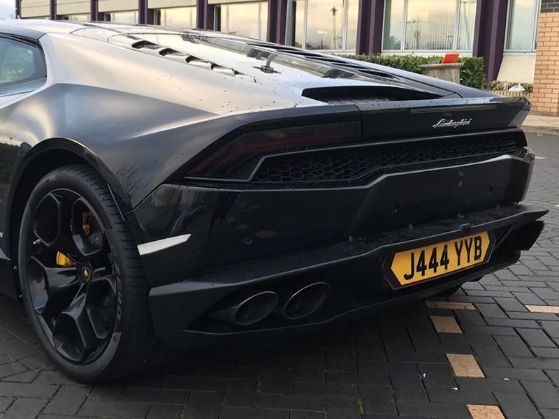 Rear three-quarter view of a black Lamborghini Huracán, featuring quad exhaust outlets, black alloy wheels and a yellow shaped number plate displaying “J444 YYB.”