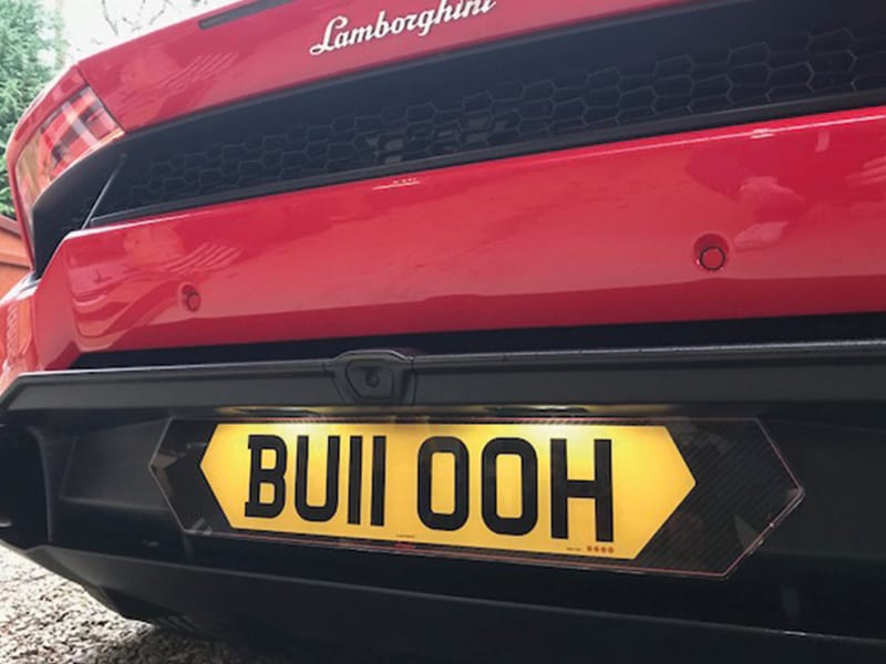Close-up rear view of a red Lamborghini Huracán, showing the Lamborghini badge and a yellow carbon fibre-effect shaped number plate displaying “BU11 OOH.”