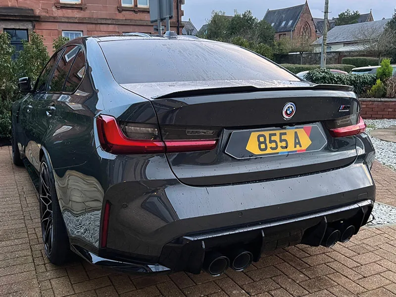 Rear three-quarter view of a grey BMW M3 G80 featuring quad exhausts, rear spoiler and a yellow M Sport-themed shaped number plate displaying “855 A.”