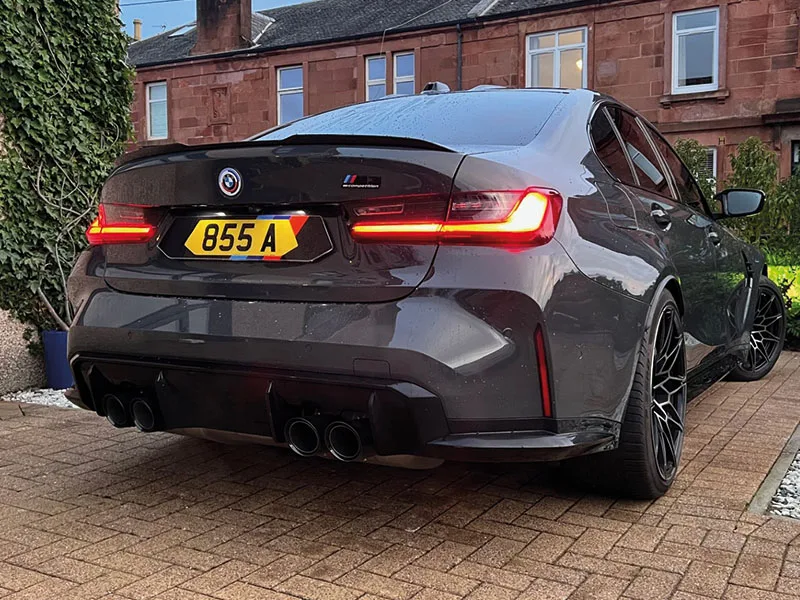 Low rear three-quarter view of a grey BMW M3 G80 showing the rear diffuser, quad exhausts and yellow shaped “855 A” number plate with BMW M-style colour detailing.