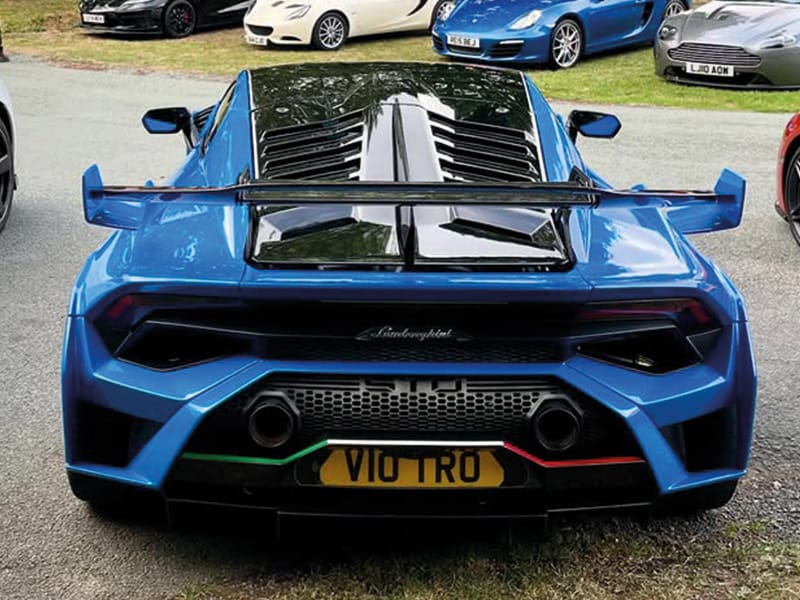 Rear view of a blue Lamborghini Huracán STO, featuring a large fixed rear wing, twin high-mounted exhausts and a yellow shaped number plate displaying “V10 TRO.”