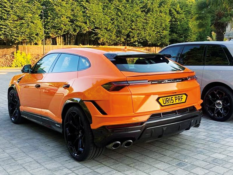 Rear three-quarter view of an orange Lamborghini Urus, showing black alloy wheels, quad exhausts and a yellow shaped number plate displaying “UR15 PRF.”