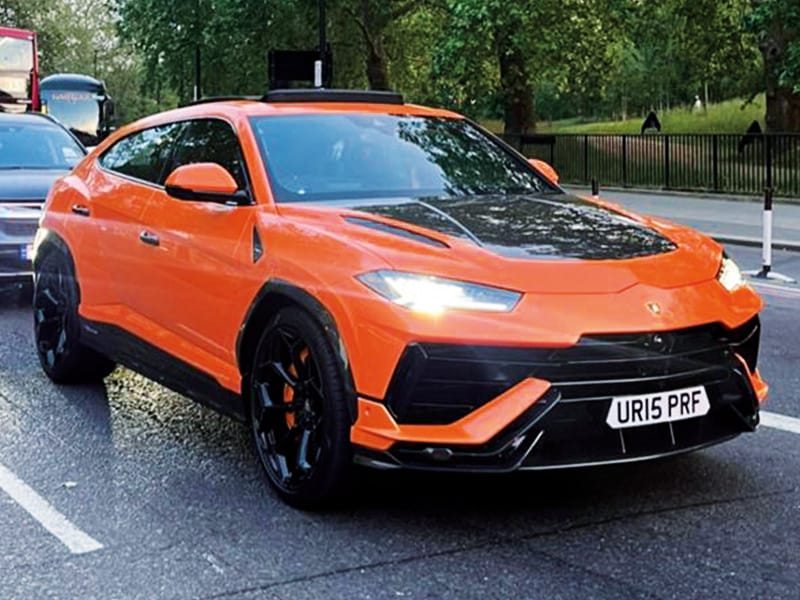 Front three-quarter view of an orange Lamborghini Urus on the road, featuring black styling details, illuminated headlights and a compact white shaped number plate displaying “UR15 PRF.