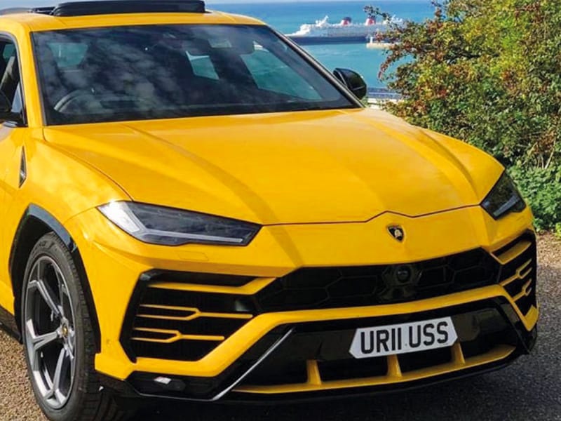 Front three-quarter view of a yellow Lamborghini Urus, showing its angular front bumper and a compact white shaped number plate displaying “UR11 USS.”