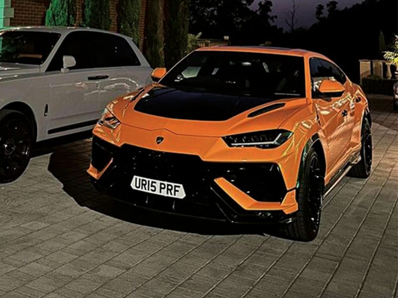Front three-quarter view of an orange Lamborghini Urus, featuring black exterior detailing and a compact white shaped number plate displaying “UR15 PRF.”