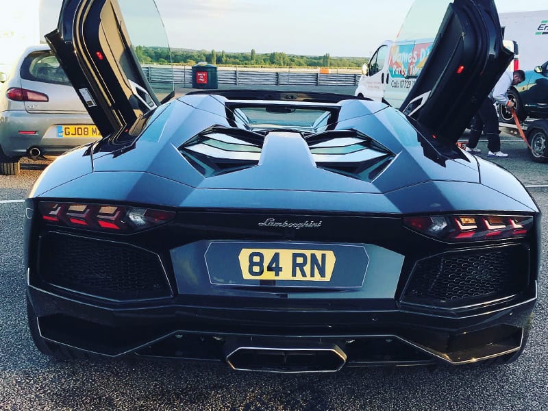 Rear view of a black Lamborghini Aventador Roadster with both scissor doors open, featuring angular taillights, a central exhaust and a yellow shaped number plate displaying “84 RN.