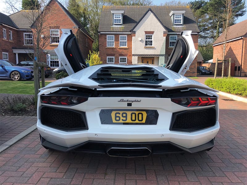 Rear view of a white Lamborghini Aventador Roadster with both scissor doors raised, showing the rear grille, angular taillights and yellow shaped number plate “69 OO.”