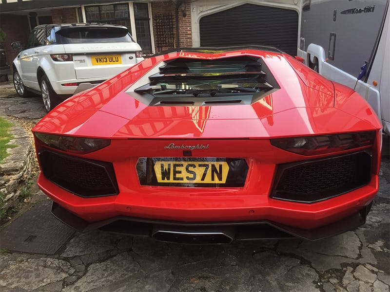 Rear view of a red Lamborghini Aventador Roadster, featuring the engine cover, black rear grilles, central exhaust and yellow shaped number plate displaying “WES7N.”