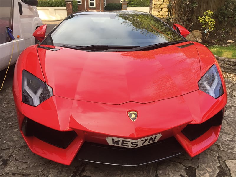 Front view of a red Lamborghini Aventador Roadster, showing its angular headlights, large front air intakes and a white shaped front number plate displaying “WES7N.
