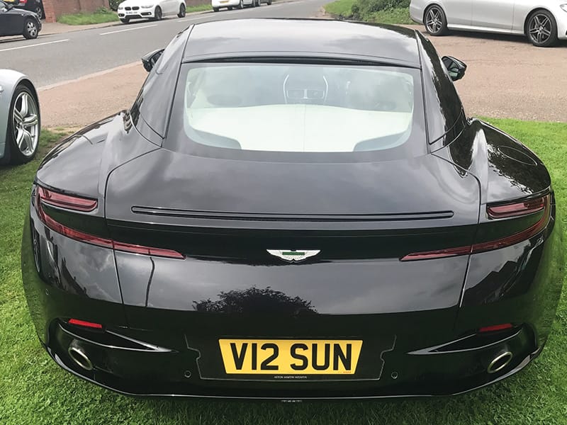 Rear view from above of a black Aston Martin DB11 parked on grass, showing the slim taillights, twin exhaust outlets and yellow UK number plate “V12 SUN.”