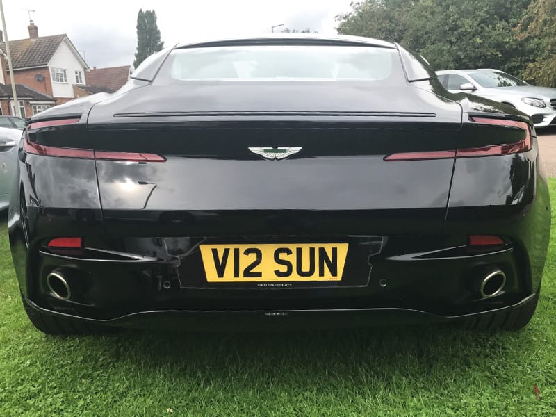 Straight rear view of a black Aston Martin DB11, featuring the sculpted rear bodywork, Aston Martin badge, twin exhausts and yellow “V12 SUN” number plate