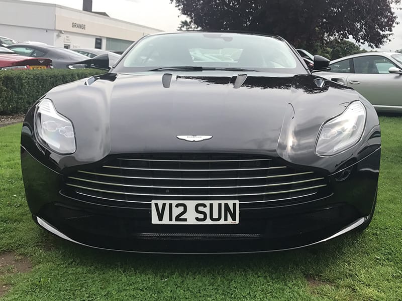 Front view of a black Aston Martin DB11 parked on grass, showing its wide grille, LED headlights and white front number plate “V12 SUN.”