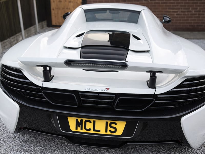 Close-up rear view of a white McLaren 650S, featuring the raised rear spoiler, twin exhaust outlets and a yellow shaped number plate displaying “MCL 1S.”