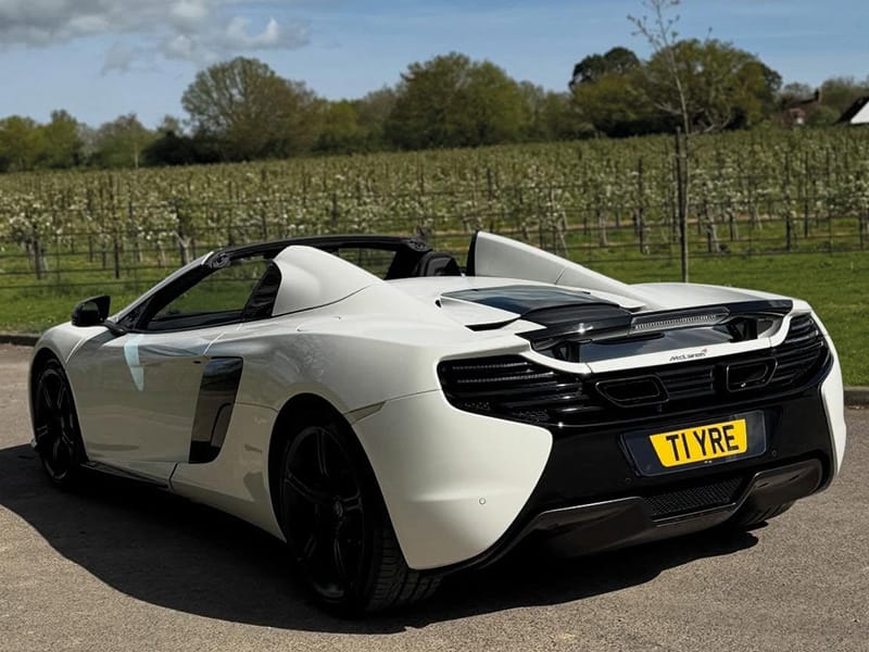 Rear three-quarter view of a white McLaren 650S Spider, showing the raised rear spoiler, black rear bodywork and a yellow shaped number plate displaying “T1 YRE.”