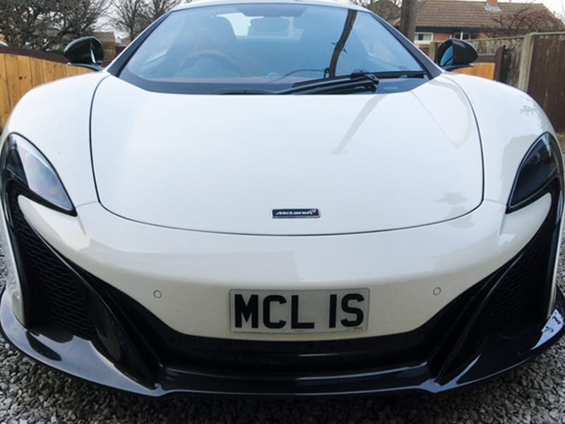 Front view of a white McLaren 650S, showing the distinctive headlight design, large front air intakes and a compact white number plate displaying “MCL 1S.”