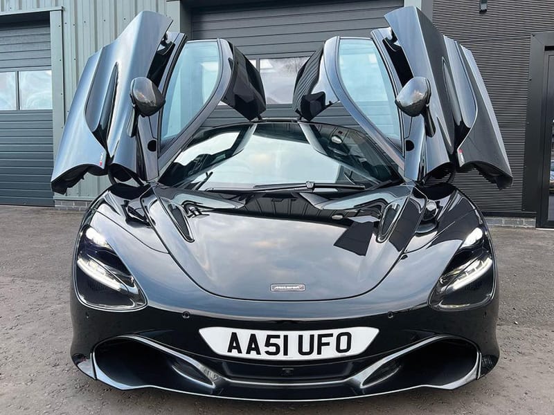 Front view of a black McLaren 720S with both dihedral doors raised, showing LED headlights and a white shaped number plate displaying “AA51 UFO.”