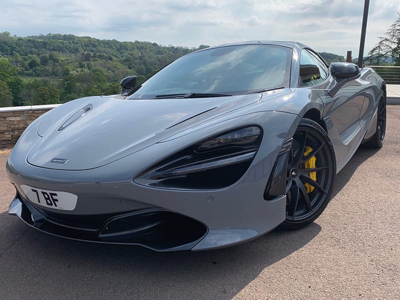 Front three-quarter view of a grey McLaren 720S, featuring black alloy wheels, yellow brake calipers and a compact white shaped number plate displaying “7 BF.”