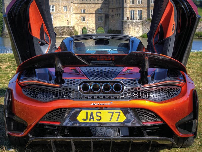 Rear view of an orange McLaren 765LT Spider with both dihedral doors raised, showing the large rear wing, quad exhausts and yellow shaped number plate displaying “JAS 77.”
