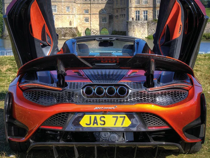 Rear view of an orange McLaren 765LT Spider with both dihedral doors raised, showing the large rear wing, quad exhausts and yellow shaped number plate displaying “JAS 77.”