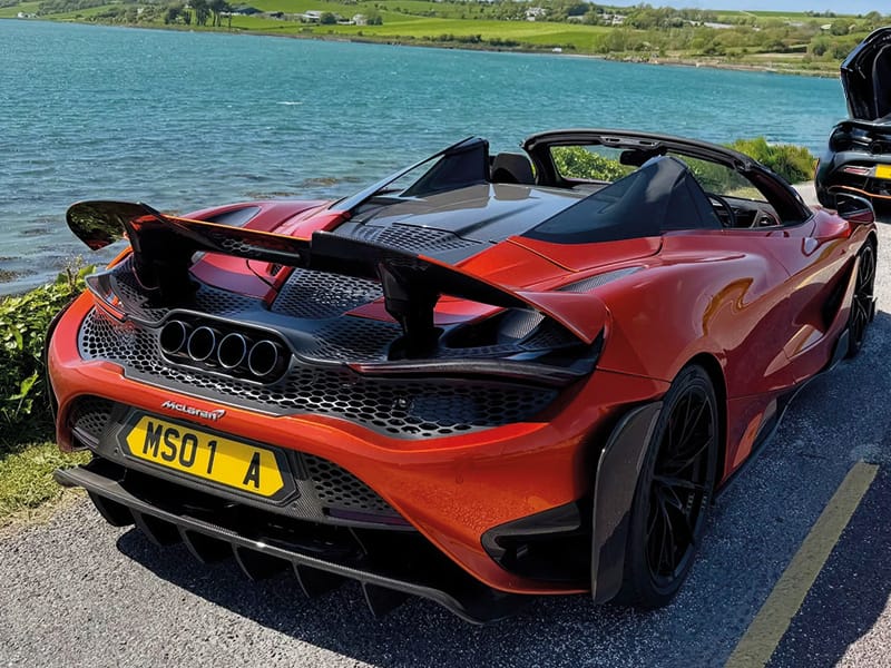 Rear three-quarter view of an orange McLaren 765LT Spider parked beside a lake, featuring the raised rear wing, quad exhausts and yellow shaped number plate displaying “MSO 1 A.”