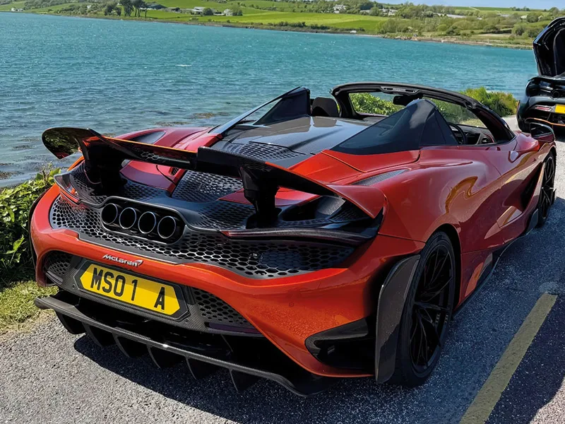 Rear three-quarter view of an orange McLaren 765LT Spider parked beside a lake, featuring the raised rear wing, quad exhausts and yellow shaped number plate displaying “MSO 1 A.”