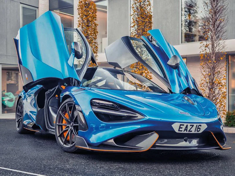Front three-quarter view of a blue McLaren 765LT with both dihedral doors raised, showing carbon fibre trim, orange accents and a white shaped number plate displaying “EAZ16.”