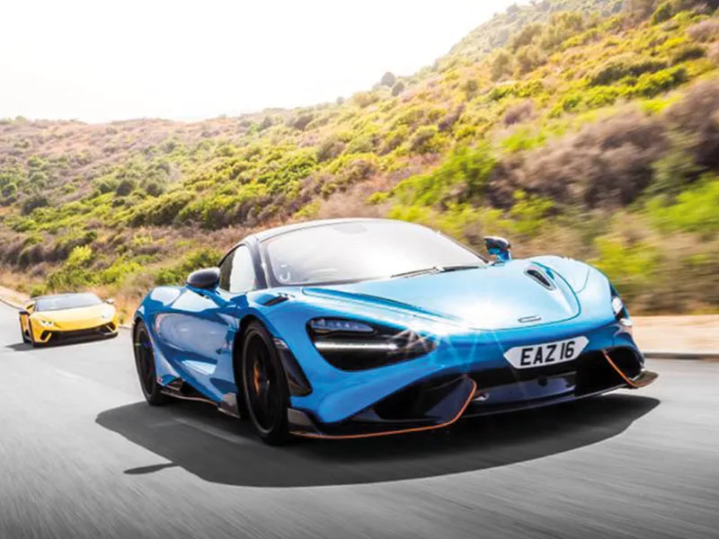 Front three-quarter view of a blue McLaren 765LT driving on a winding road, featuring carbon fibre aerodynamic styling and a white shaped number plate displaying “EAZ16.”