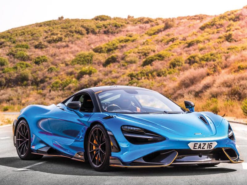 Front three-quarter view of a blue McLaren 765LT parked on a road, showing black alloy wheels, carbon fibre detailing and a white shaped number plate displaying “EAZ16.”