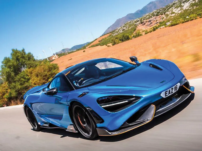 Front three-quarter view of a blue McLaren 765LT driving on a mountain road, featuring carbon fibre aerodynamic details and a white shaped number plate displaying “EAZ16.”