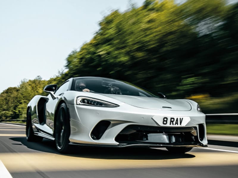 Front three-quarter view of a white McLaren GT driving on the road, featuring black aerodynamic trim and a white shaped number plate displaying “8 RAV.”