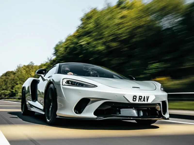 Front three-quarter view of a white McLaren GT driving on the road, featuring black aerodynamic trim and a white shaped number plate displaying “8 RAV.”