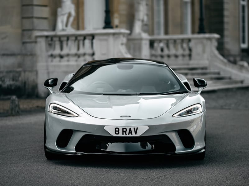 Straight front view of a white McLaren GT, showing slim headlights, large front air intakes and a white shaped number plate displaying “8 RAV.”
