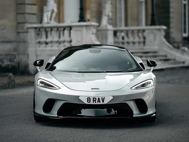 Straight front view of a white McLaren GT, showing slim headlights, large front air intakes and a white shaped number plate displaying “8 RAV.”