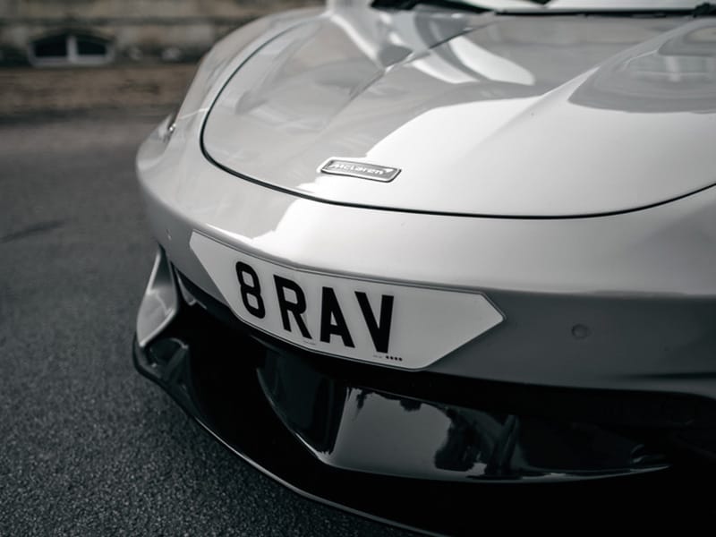 Close-up front view of a white McLaren GT, highlighting the bonnet badge, black lower grille and white shaped number plate displaying “8 RAV.”