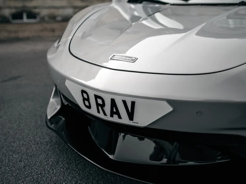 Close-up front view of a white McLaren GT, highlighting the bonnet badge, black lower grille and white shaped number plate displaying “8 RAV.”