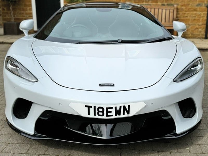 Front view of a white McLaren GT, showing its sculpted bumper, black air intakes and a white shaped number plate displaying “T18 EWN.”
