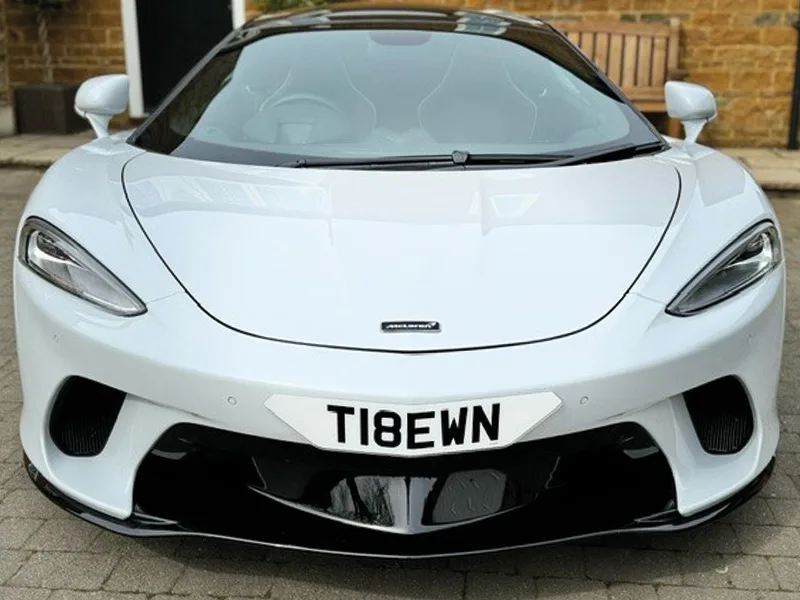 Front view of a white McLaren GT, showing its sculpted bumper, black air intakes and a white shaped number plate displaying “T18 EWN.”