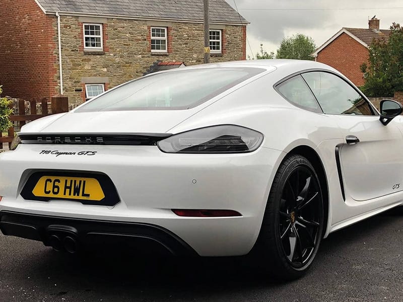 Rear three-quarter view of a white Porsche 718 Cayman GTS, showing black alloy wheels, central twin exhausts and a yellow black-surround shaped number plate displaying “C6 HWL.”