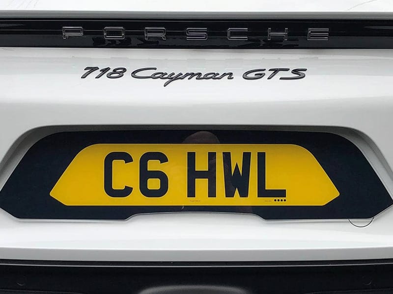 Close-up rear view of a white Porsche 718 Cayman GTS, showing Porsche and 718 Cayman GTS badging with a yellow shaped number plate displaying “C6 HWL.”