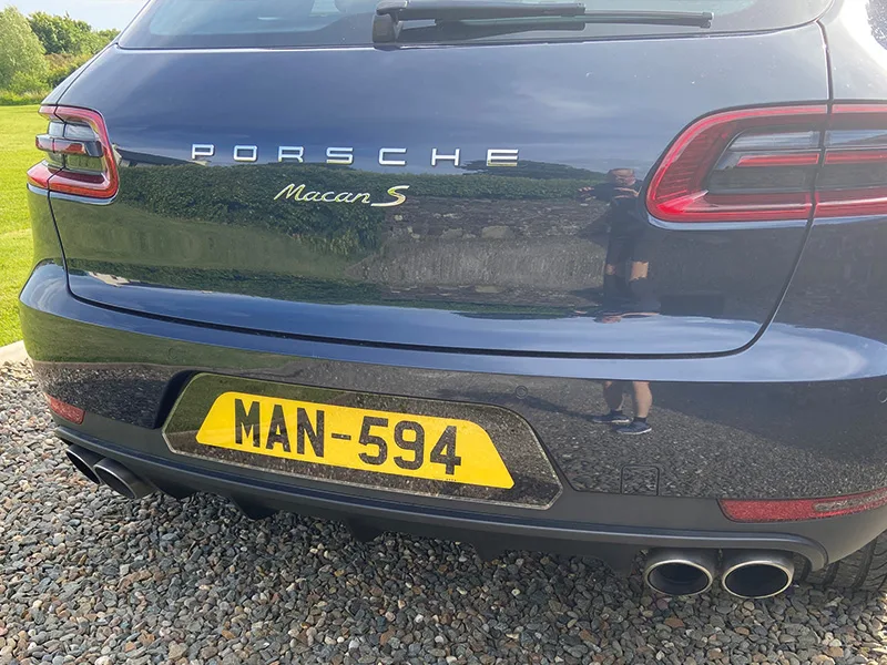 Close-up rear view of a black Porsche Macan S, showing Porsche and Macan S badging, quad exhausts and a yellow shaped number plate displaying “MAN-594.”