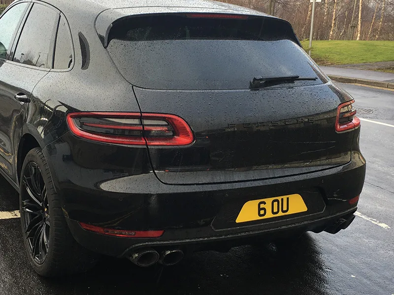 Rear three-quarter view of a black Porsche Macan, featuring LED taillights, quad exhaust outlets and a yellow black-surround shaped number plate displaying “6 OU.”