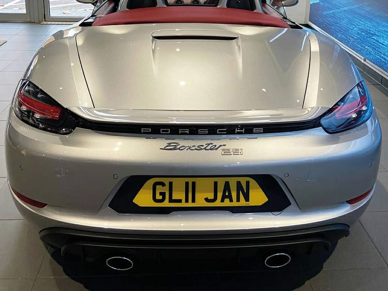 Rear view of a silver Porsche 718 Boxster 25, featuring Porsche and Boxster 25 badging, twin exhaust outlets and a yellow black-surround shaped number plate displaying “GL11 JAN.”