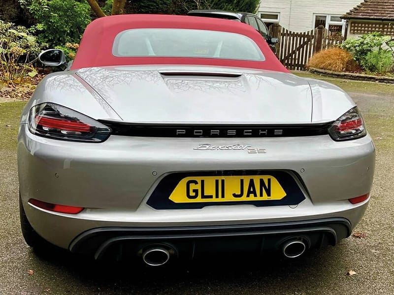 Rear view of a silver Porsche 718 Boxster 25 with a red fabric roof, showing twin exhaust outlets and a yellow black-surround shaped number plate displaying “GL11 JAN.”